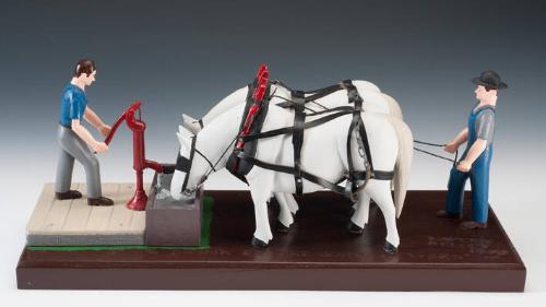 A painted wooden sculpture of two white horses in full harness drinking from a trough; a man in…