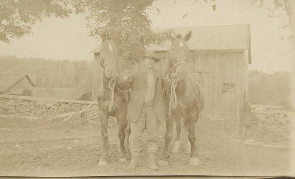 Man Holding Two Horses In Front of Barn