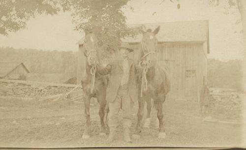 Man Holding Two Horses In Front of Barn